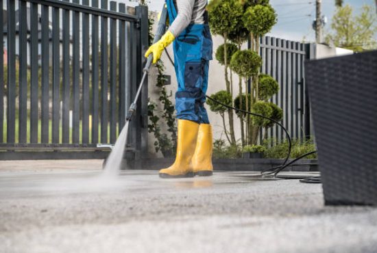 istockphoto-1418464469-612×612 Man in Yellow Rain Boots and Work Uniform Pressure Washing Concrete Tiles of the Driveway to His House Behind the Closed Yard Entrance Gate. Home Surroundings Maintenance Theme.